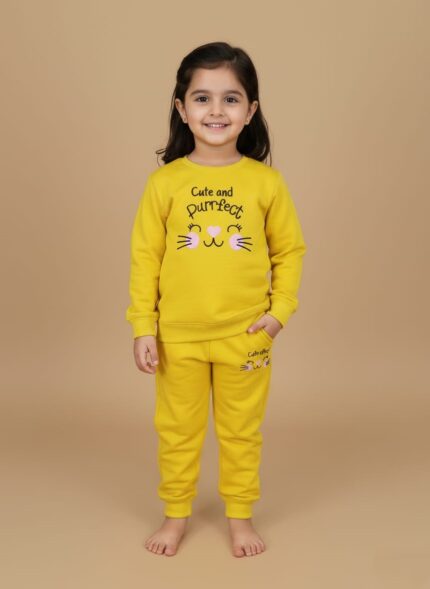 Full-length image of a smiling young girl wearing a bright yellow Girls Yellow Tracksuit set, consisting of a "Cute and Purrfect" cat-themed sweatshirt and matching joggers, standing barefoot against a beige background.