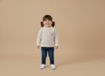Smiling toddler girl in a light pastel knit sweater with a cute animal applique, blue jeans, and white canvas shoes, standing on a beige background.