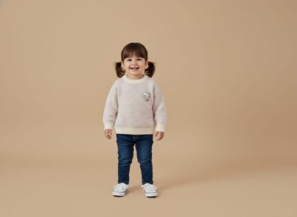 Smiling toddler girl in a light pastel knit sweater with a cute animal applique, blue jeans, and white canvas shoes, standing on a beige background.